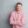 Young redhead man over grey grunge wall wearing pink shirt happy face smiling with crossed arms looking at the camera. Positive person.