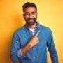 Young indian man wearing denim shirt standing over isolated yellow background cheerful with a smile of face pointing with hand and finger up to the side with happy and natural expression on face